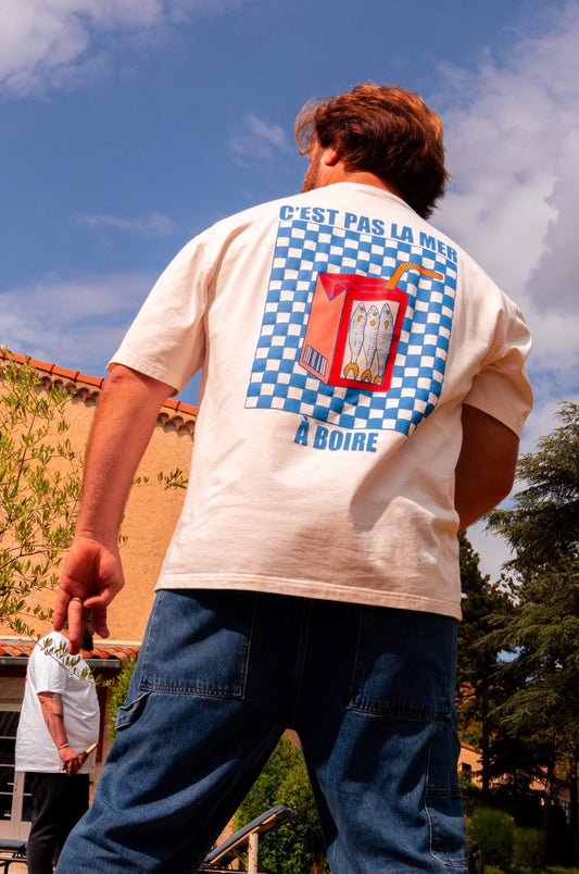 T-shirt oversize beige avec un damier bleu dans le dos et une brique avec trois poissons à l'intérieur.